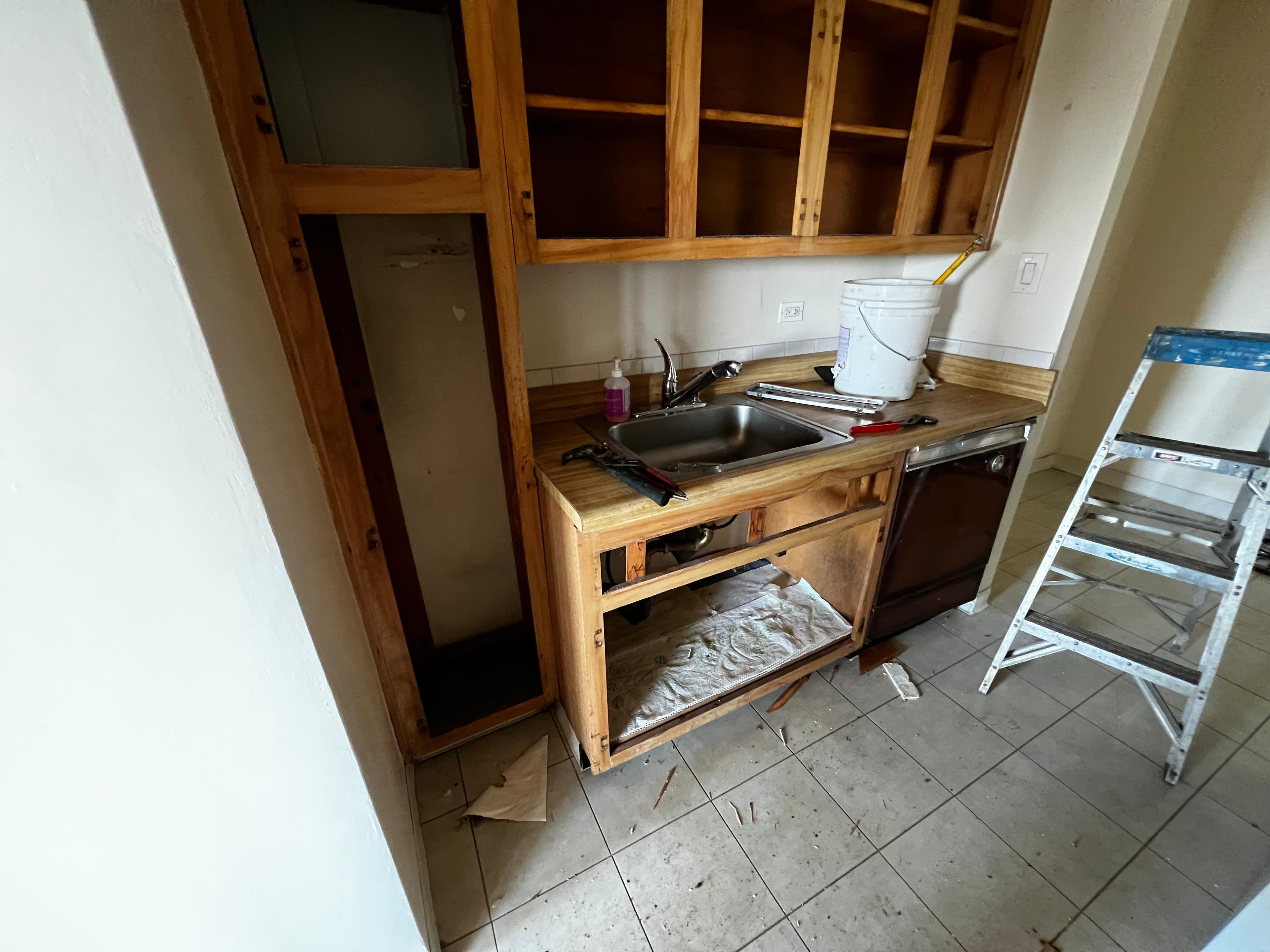 Kitchen renovation featuring wooden cabinets, a stainless steel sink, tools, and a step ladder.