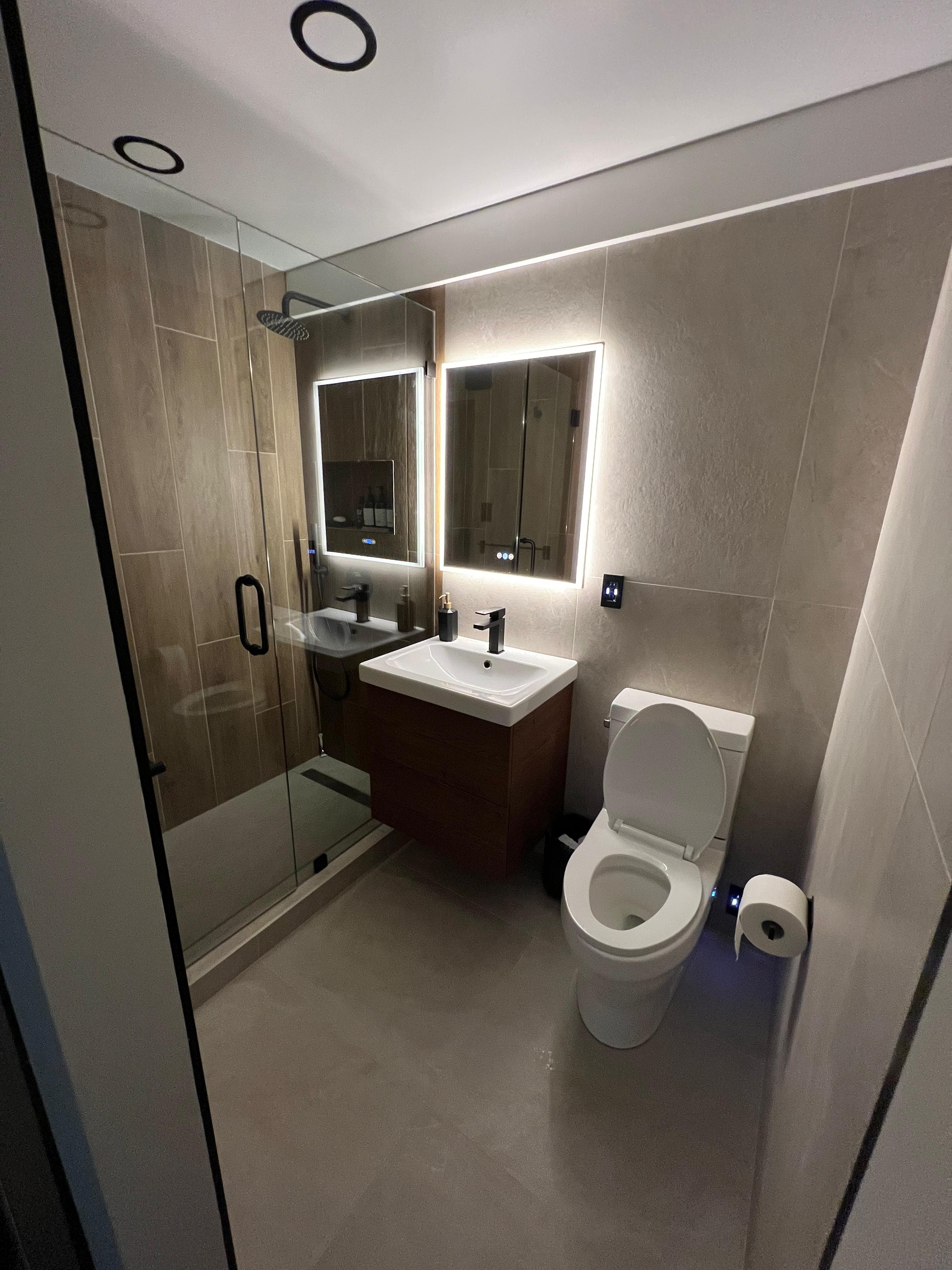 Modern bathroom featuring a glass shower, wood vanity with backlit mirror, and white toilet.